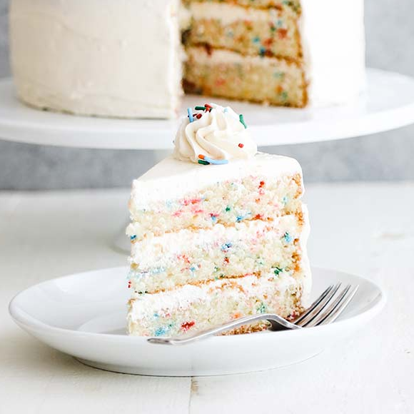 a vanilla confetti layer cake with white icing, on a white plate with a fork next to it